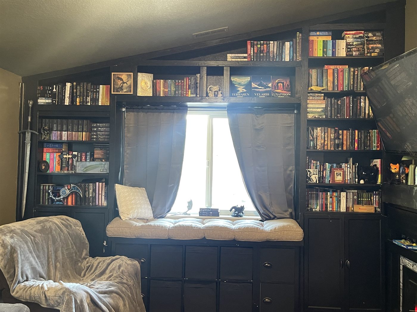 A hand-built black floor-to-ceiling home library framing a window reading nook with a cushioned bench, blackout curtains, book-filled shelves, a cozy recliner, swords and figurines, and a painted TERRASEN VELARIS LUNATHION sign