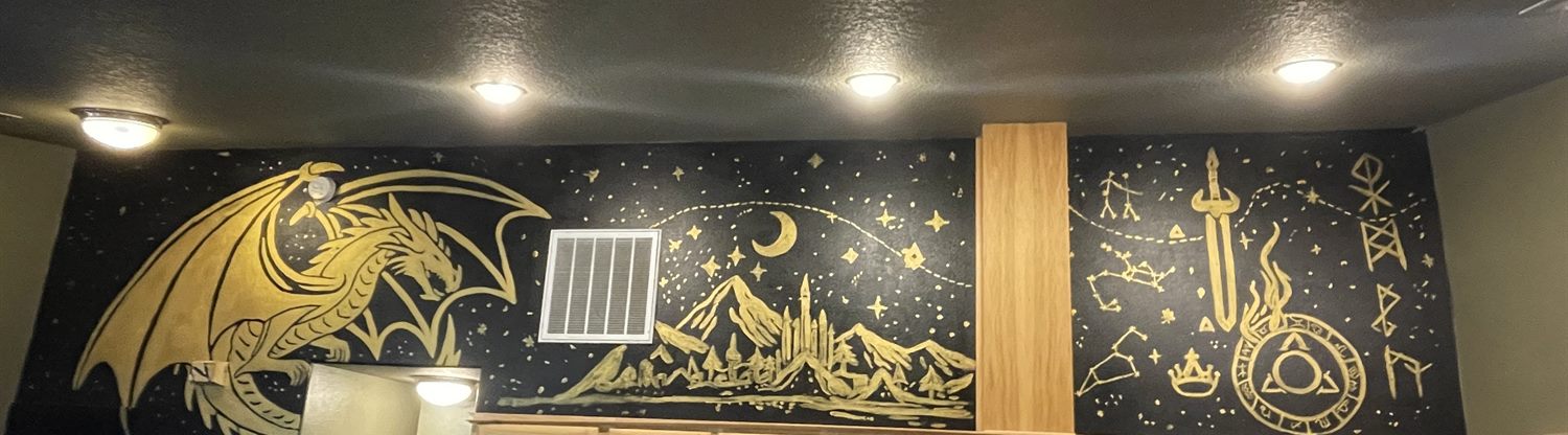 A black accent wall hand-painted in gold with a dragon, mountains and a castle, a crescent moon and stars, a sword wreathed in flame, constellations and runes — the mural that inspired the MOBS black-and-gold color scheme
